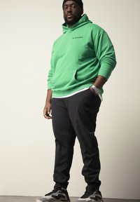 Groene hoodie met een voorzak en "STHUGE" logo, gecombineerd met zwarte broek en zwart-grijze sneakers. Eenvoudig ontwerp, casual fit.