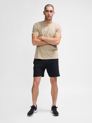 Ung mand står med armene over kors og har beige kortærmet skjorte, sorte shorts og sorte sneakers på mod en hvid baggrund.