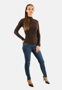 Maglione a collo alto a coste marrone abbinato a jeans skinny in denim scuro e tacchi a spillo neri. La modella è in posa con una mano sui fianchi.