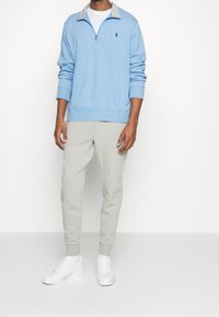 Ljusblå zip-up sweatshirt med grå krage, broderad logotyp på bröstet, kombinerad med ljusgrå avsmalnande mjukisbyxor och vita sneakers.