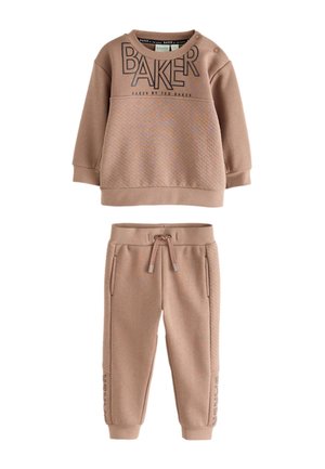 Ung dreng i beige matchende sweatshirt og bukser med hvide sneakers, der står på et lyst gulv mod en ensfarvet væg.