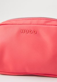 Pochette rose corail en matériau synthétique texturé, avec une fermeture éclair argentée et le logo "HUGO" en relief sur le devant. Design compact et élégant.