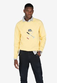 Sudadera amarilla con un gráfico que presenta una gorra y herramientas cruzadas. Mangas largas, cuello redondo, llevada sobre una camisa a rayas. Pantalones negros.