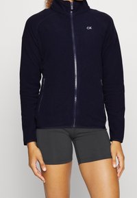 Marinblå fleecejacka med hög krage, full dragkedja fram och två sidfickor. Har en liten logotyp på övre vänstra sidan.