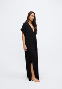 Fekete maxi ruha rövid ujjakkal, mély V-nyakkivágással és fém díszítéssel a nyakkivágásnál. Oldalsó hasítékokkal a nagyobb mozgékonyság és áramlás érdekében.