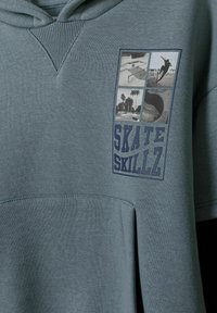 Sudadera en un suave color teal con un bolsillo canguro. Presenta un parche gráfico con imágenes de skateboard y el texto "SKATE SKILLZ" en azul.
