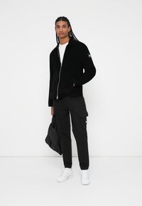 Svart zip-up jakke laget av mykt materiale, brukt over en hvit t-skjorte. Kombinert med sorte cargosbukser og hvite joggesko. Minimalistisk design.
