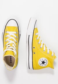Gule høje sneakers i lærred med hvide snørebånd, gummitåhætte, sort og hvid stribe på sålen og Converse-logo.