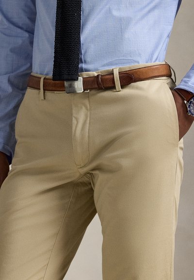 Man draagt khaki broek, bruine leren riem, blauw geruit overhemd, zwarte gestructureerde stropdas en een blauw horloge met bruine band.