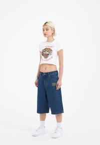 White cropped t-shirt with tiger graphic and red text paired with blue denim culottes, featuring yellow embroidered detail and white sneakers.