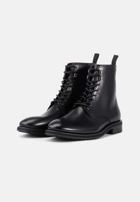 Botoșei negri din piele, cu o finisare netedă, cu vârf rotund, sistem de șireturi și branțuri laterale. Talpă groasă din cauciuc, cu un model texturat.