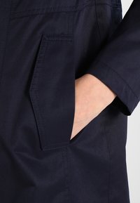 Cappotto blu navy con tasca laterale visibile, texture liscia e leggera lucentezza. Il design include dettagli cuciti lungo le cuciture.