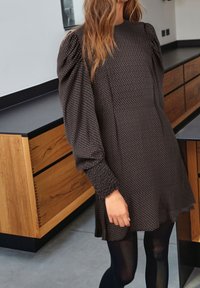 Tummalla kuviollisella mekolla, jossa on puffihihat ja rypytetyt hihansuut. Kankaassa on pieni geometrinen kuvio. Yhdistetty mustiin tiheisiin sukkahousuihin.