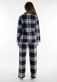 Vrouw die blootsvoets met de rug naar je toe staat, gekleed in een zwart-wit-grijze geruite pak met lange broek en een bijpassende jas.