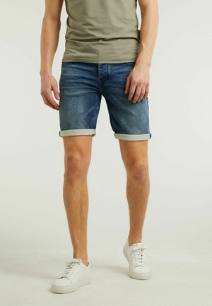 Man draagt opgerolde blauwe spijkershorts, olijfgroene T-shirt en witte sneakers, staat tegen een effen achtergrond.