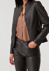 Veste en cuir noir avec col montant, fermeture zippée et finitions texturées, associée à un chemisier beige et un pantalon en cuir noir assorti.