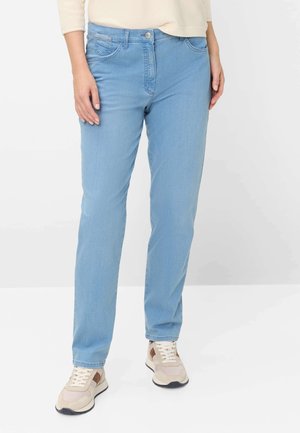 Frau trägt hellblaue Straight-Leg-Jeans, beige Sneaker und ein cremefarbenes Langarmshirt, steht auf einem weißen Boden vor weißem Hintergrund.