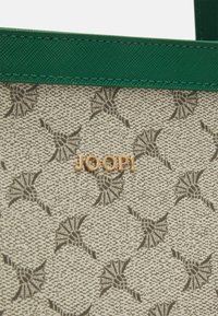 Tissu beige texturé avec un motif floral abstrait sombre et une bordure verte, avec des lettres métalliques dorées épelant JOOP! sur un sac.