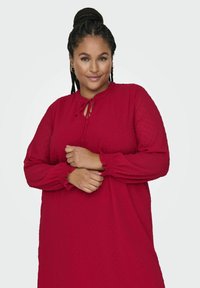 Czerwona sukienka z długim rękawem, okrągłym dekoltem i detalem w postaci wiązania. Delikatna struktura z kropkowym wzorem oraz marszczonymi mankietami. Luźny krój.