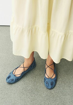 Füße tragen blaue bestickte Ballerinas mit kreuzenden Riemen, sichtbar unter einem langen, hellgelben Stufenkleid auf einem grauen Boden.