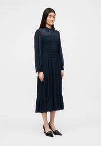 Μίντι φόρεμα σε ναυτικό μπλε με γιακά, ελαστικό μπούστο, κλείσιμο με κουμπιά και μακριά διαφανή μανίκια· διαθέτει στρωματοποιημένο τούλινο τελείωμα. Συνδυάζεται με μαύρα τακούνια.