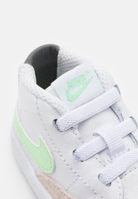 White leather sneaker with green Nike swoosh, white laces, and a grey mesh lining. Notable tan suede accent on the sole.