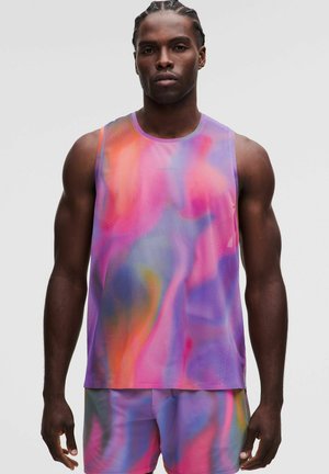 Uomo con capelli intrecciati che indossa una canotta sportiva multicolore rosa, viola e arancione e pantaloncini abbinati, in piedi davanti a uno sfondo semplice.