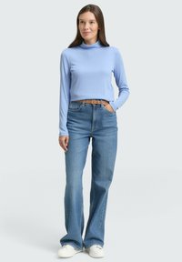 Vrouw die rechtop staat, gekleed in een lichtblauwe turtleneck, hoge taille blauwe jeans met een bruine riem en witte sneakers, tegen een effen achtergrond.