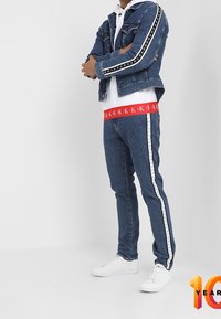 Muž v modré džínové bundě a džínách s bílým pruhem, bílém mikině, červeném pásku Calvin Klein a bílých teniskách, stojí se zkříženýma rukama.