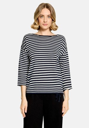 Frau mit kurzen blonden Haaren, die ein schwarz-weiß gestreiftes Langarmshirt und schwarze Hose trägt, steht vor einem einfarbigen Hintergrund.