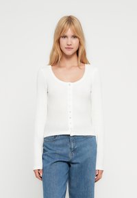 Γυναίκα με ίσια ξανθά μαλλιά, που φοράει εφαρμοστή λευκή μπλούζα με μακριά μανίκια και κουμπιά, καθώς και μπλε ψηλόμεσα τζιν, κοιτάζοντας προς τα εμπρός.