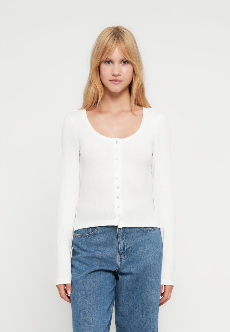 Γυναίκα με ίσια ξανθά μαλλιά, που φοράει εφαρμοστή λευκή μπλούζα με μακριά μανίκια και κουμπιά, καθώς και μπλε ψηλόμεσα τζιν, κοιτάζοντας προς τα εμπρός.