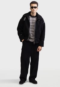 Black fleece jacket with leather accents, textured sweater with a wavy pattern, black wide-leg pants, and dark shoes. Accessories include sunglasses.