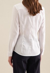 Valkoinen pitkähiha blouse, jossa on istuva siluetti, napitettu hihansuu ja hienovaraiset takapleatit. Sileä kangas, jossa on hieman kiiltoa.