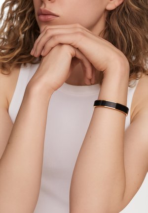 Black and gold bangle bracelet against a light-skinned arm, featuring a sleek design and smooth surface, worn on a plain white top.