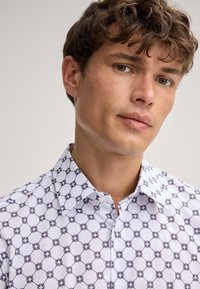 Chemise blanche avec un motif géométrique de contours foncés alternés et de cercles, dotée d'un col classique et d'une patte de boutonnage.