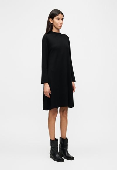 Μακρυμάνικο μαύρο φόρεμα από απαλό ύφασμα, με ψηλό λαιμόκοψη και σιλουέτα Α γραμμής. Συνδυάζεται με μαύρα μποτάκια μέχρι τον αστράγαλο.