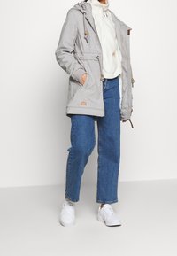 Gray hooded jacket with drawstrings, featuring a zip front and pockets, worn over a cream sweater and blue jeans, paired with white sneakers.