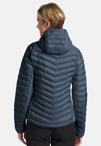 Femme aux cheveux blonds courts portant une veste à capuche bleu matelassée et un pantalon noir, vue de dos.