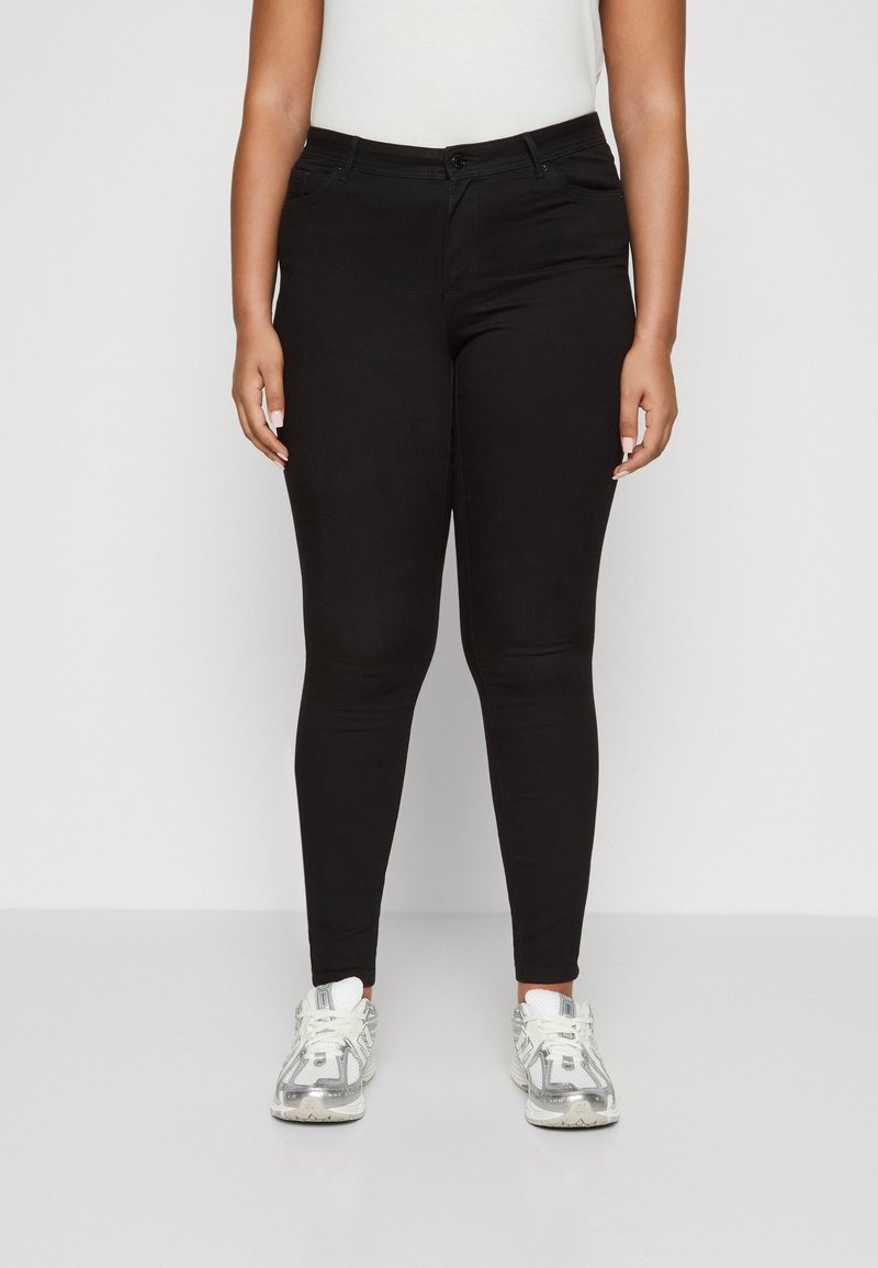 Sorte tettsittende jeans med en glatt tekstur, som har knapplås og glidelås, frontlommer og en tettsittende design. Kombinert med hvite sneakers.