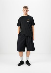 Ung mann står mot hvit bakgrunn iført svart T-skjorte med kenguru-logo, løse svarte shorts, hvite sokker og svarte joggesko.