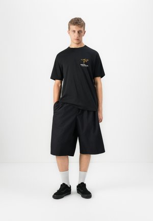 Ung mann står mot hvit bakgrunn iført svart T-skjorte med kenguru-logo, løse svarte shorts, hvite sokker og svarte joggesko.