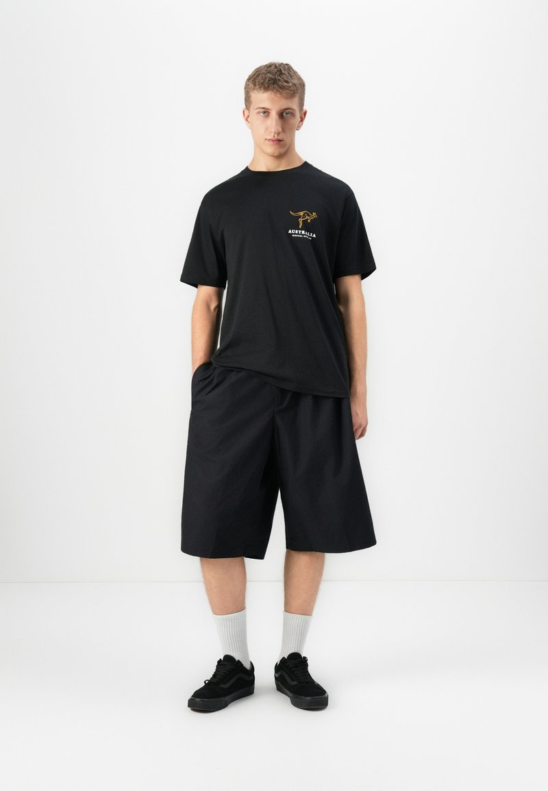 Ung mann står mot hvit bakgrunn iført svart T-skjorte med kenguru-logo, løse svarte shorts, hvite sokker og svarte joggesko.