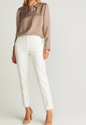 Woman wearing taupe satin long-sleeve blouse, white tailored ankle pants, and beige high heels, standing against a plain background.