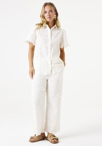 Witte, lichte, geborduurde katoenen blouse met korte mouwen en knopen aan de voorkant, gecombineerd met bijpassende palazzo-broeken. Bruine sandalen.