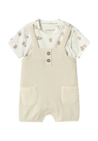 Peto de color beige con relieve, con dos bolsillos y cierre de botones. Debajo lleva una camiseta blanca con un estampado de animales marrones.