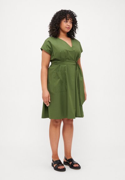 Abito verde oliva con design avvolgente, maniche a cappuccio, due tasche frontali e cintura in vita. Abbinato a sandali neri. Tessuto dalla superficie liscia.