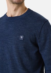 Marinblauwe gebreide trui met een ronde halslijn en geribbelde boorden, met een klein logo-op-patch op de borst en een textuurpatroon in de stof.