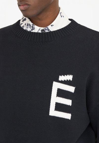 Pull noir en tricot avec un logo « É » blanc sur le devant, présentant une finition texturée et un col côtelé, superposé à une chemise blanche à motifs floraux.