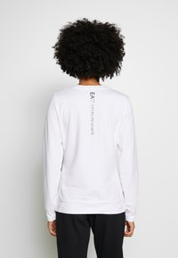 Vit långärmad skjorta i bomull med rund hals och ett vertikalt svart logotryck längs mitt bak. avslappnad passform.